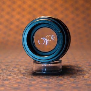 Puede incluir: Un objetivo de c&aacute;mara negro con un centro de cristal transparente, apoyado sobre un soporte cil&iacute;ndrico transparente. La lente tiene un tono azul-verdoso y el texto "Nikon" es visible. El fondo es naranja y marr&oacute;n estampado.