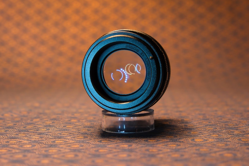 Puede incluir: Un objetivo de c&aacute;mara negro con un centro de cristal transparente, apoyado sobre un soporte cil&iacute;ndrico transparente. La lente tiene un tono azul-verdoso y el texto "Nikon" es visible. El fondo es naranja y marr&oacute;n estampado.