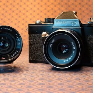 May include: A black Praktica LLC camera with a lens and a separate Pentacon lens. The lenses have text that reads "electric", "MADE IN G.O.R.", and "aus JENA DOR". The camera is on a patterned surface.