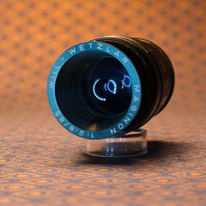 Puede incluir: Un objetivo de c&aacute;mara vintage con el texto "WILL-WETZLAR MAGINON 1:2.8/85" en letras blancas sobre un anillo azul verdoso. El objetivo es negro con una base transparente, sobre un fondo naranja estampado. Un accesorio de fotograf&iacute;a.