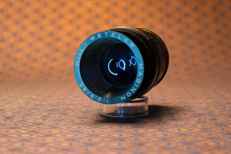 Puede incluir: Un objetivo de c&aacute;mara vintage con el texto "WILL-WETZLAR MAGINON 1:2.8/85" en letras blancas sobre un anillo azul verdoso. El objetivo es negro con una base transparente, sobre un fondo naranja estampado. Un accesorio de fotograf&iacute;a.