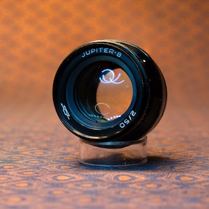 May include: A black Jupiter-8 2/50 camera lens, resting on a clear stand. The lens has the text "JUPITER-8" and "2/50" printed on it. The background is a patterned orange and blue surface.