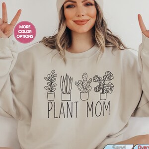 Könnte beinhalten: Ein beiger Pullover mit einer Grafik von Topfpflanzen und dem Text "PLANT MOM".