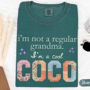 以下が含まれることがあります： 「I'm not a regular grandma. I'm a cool COCO」の文字が入ったブルーグリーンのTシャツ。「COCO」の文字はパッチワーク生地で作られています。コーヒーカップ、サングラス、ノートも画像に写っています。