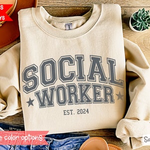 May include: A beige sweatshirt with the text "SOCIAL WORKER EST. 2024" in gray lettering with a star on each side of the word "WORKER".