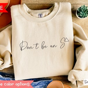 Może przedstawiać: Beżowa bluza z napisem "Don't be an S" w czarnym stylu pisma odręcznego z symbolem trójkąta. Bluza leży płasko na drewnianej powierzchni.
