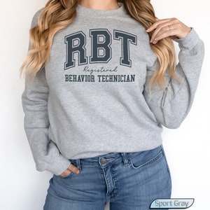 May include: A gray sweatshirt with the text "RBT Registered Behavior Technician" in black lettering. The sweatshirt is being worn by a person with long blonde hair.