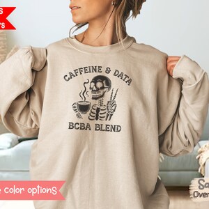 Pode incluir: Camisola oversized cor de areia com o texto "Caffeine & Data BCBA Blend" e um gráfico de um esqueleto segurando uma xícara de café e fazendo o sinal de paz. A camisola é de cor neutra.