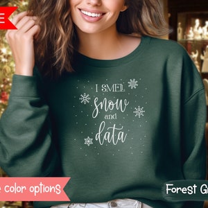 Op de afbeelding: Bostgroen sweatshirt met de witte tekst "I smell snow and data" en sneeuwvlokaccenten. De sweatshirt heeft een ronde hals en lange mouwen. Het woord "SALE" staat in een rode banner linksboven.