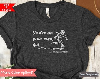 May include: Dark grey heather t-shirt with the text "You're on your own, kid." and a running figure in white. The phrase "You always have been" is below the figure. The shirt is a crew neck.