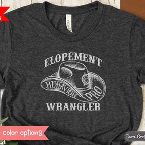 Peut inclure: T-shirt gris foncé avec un graphique blanc d'un chapeau de cowboy et le texte "ELOPEMENT BEHAVIOR PRO WRANGLER".
