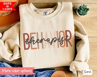 May include: A beige sweatshirt with the text "BEHAVIOR therapist" printed in black and brown on the front. The text is layered with different shades of brown, creating a gradient effect.