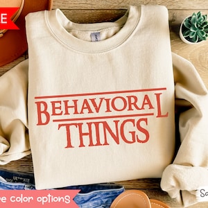 Puede incluir: Una sudadera beige con el texto "BEHAVIORAL THINGS" en rojo, combinada con un sombrero marrón y unos vaqueros azules. La sudadera está disponible en el color "Arena".