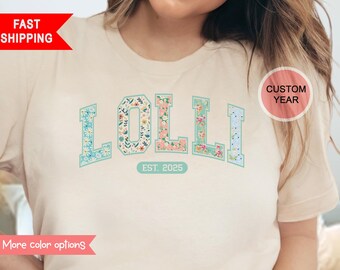 May include: A light beige t-shirt with the word "LOLLI" in large, teal letters with a floral pattern. The letters are outlined in teal and the year "EST. 2025" is printed below the word.