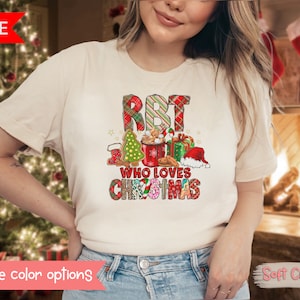 Op de afbeelding: Crèmekleurig T-shirt met een kerstthema. De afbeelding bevat de tekst "RBT WHO LOVES CHRISTMAS" in een geruit patroon, samen met peperkoek, een kerstboom en een kerstmuts. Het shirt wordt door een persoon gedragen.