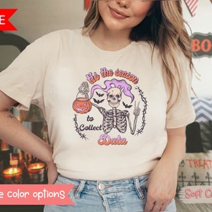 Pode incluir: Uma camiseta cor creme com um gráfico de um esqueleto segurando uma abóbora e o texto "Tis the season to collect data".