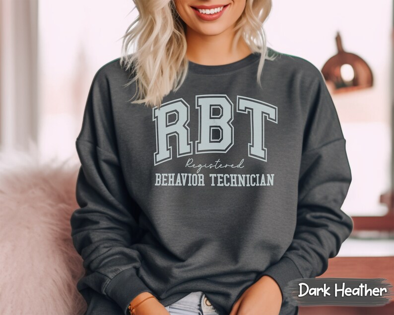 May include: Dark heather grey sweatshirt with the text "RBT Registered Behavior Technician" in a light blue color with a varsity style font.