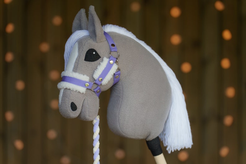 May include: A gray and white stuffed horse head with a purple halter and a white mane. The horse is on a wooden stick with a white and purple rope attached.