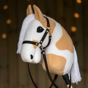 May include: A plush hobby horse with a white and tan spotted pattern. It features a black bridle with silver-colored hardware and a white mane and tail. The toy is mounted on a wooden stick.