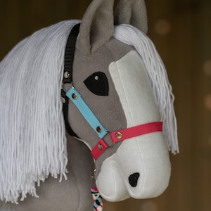 Peut inclure: Une tête de cheval à bascule en peluche grise et blanche avec une crinière blanche. Le cheval a un licol noir avec une sangle bleue et rose. Une corde multicolore est attachée au licol. Le jouet est conçu pour être monté sur un bâton.