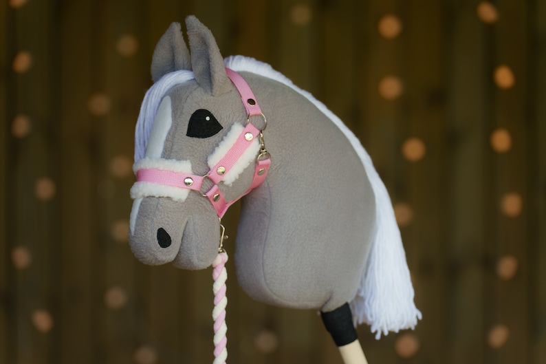May include: A gray and white stuffed horse head with a pink bridle and white mane. The horse is attached to a wooden stick.