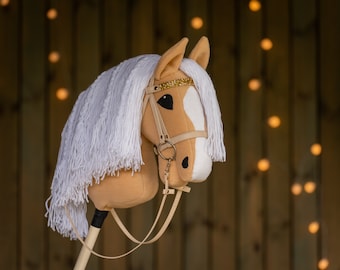 May include: A tan and white stuffed horse head with a white mane and tail. The horse has a brown leather bridle with a gold trim. The horse is on a wooden stick.