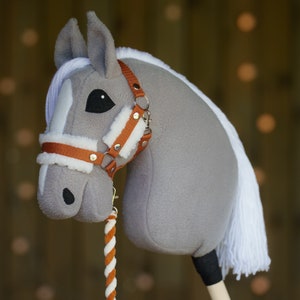 May include: A gray and white stuffed horse head with a brown leather halter and a white and orange rope rein. The horse has a black eye and a white mane.