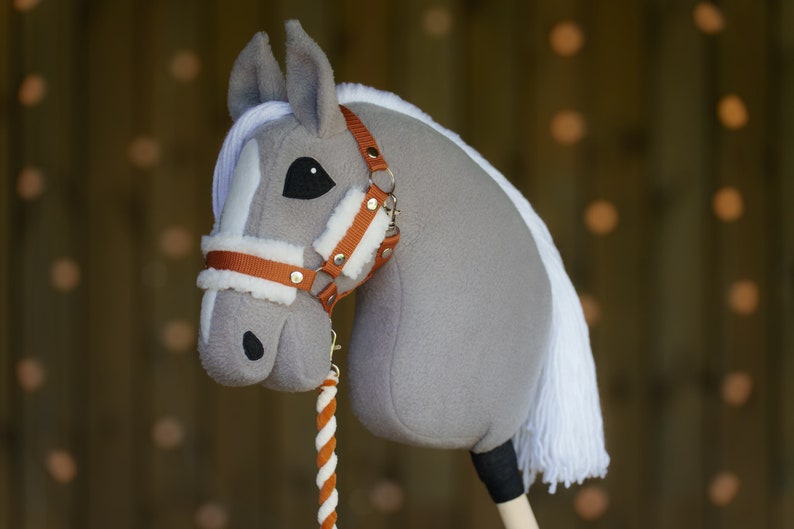 May include: A gray and white stuffed horse head with a brown leather halter and a white and orange rope rein. The horse has a black eye and a white mane.