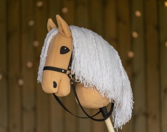 May include: A tan hobby horse with a white mane and tail. The toy horse has black eyes and a black bridle. The horse is mounted on a wooden stick.