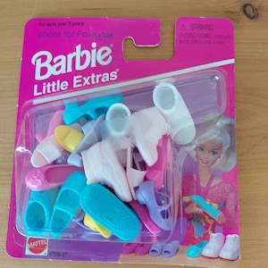 May include: A package of Barbie Little Extras shoes for poseable dolls. The package is pink and clear, with a variety of colorful doll shoes, including sandals, heels, and boots. The package also features the Barbie logo and a warning about small parts.