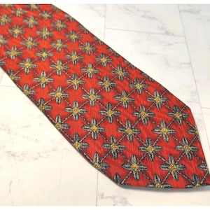 Puede incluir: Una corbata roja con un patrón repetido de diseños florales amarillos y grises. La corbata está hecha de una tela texturizada y está en ángulo sobre una superficie blanca.