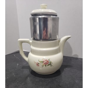 May include: A cream-colored ceramic teapot with a floral pattern featuring pink roses and green leaves. The teapot has a metallic silver-colored cylindrical insert at the top, topped with a matching cream-colored lid with a small knob handle.