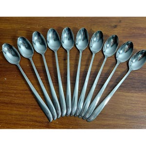 May include: A set of ten matching stainless steel long-handled spoons arranged in a fan shape. Each spoon features a polished silver finish and a slender handle with a small, engraved starburst or floral detail near the base of the stem.