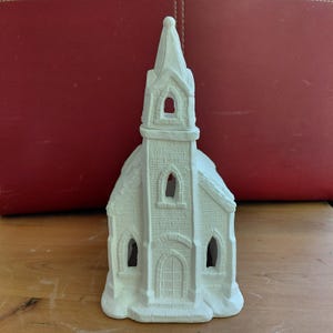 May include: A white ceramic church figurine featuring a tall steeple, arched windows, and a textured brick pattern on the exterior walls. The structure includes a central arched doorway and open window cutouts, presented in a matte finish.