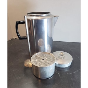 May include: A vintage aluminum coffee percolator with a black handle, disassembled. The components include the main pot, a filter basket, a lid with a glass knob, and a stem. The percolator is designed for stovetop use.