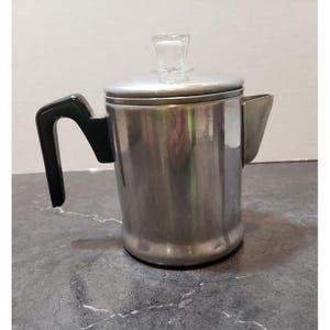 May include: A stainless steel coffee percolator with a black handle and a glass knob on the lid. The percolator has a spout and is sitting on a gray surface. The coffee pot is designed for brewing coffee.