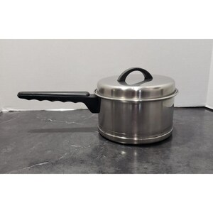 May include: Stainless steel saucepan with a lid and a black handle. The pot has a black handle and a black handle on the lid. The pot is sitting on a dark gray surface.