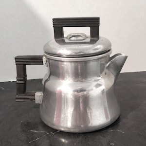 May include: A silver-colored aluminum stovetop coffee percolator with a wide, rounded base and a short, angled spout. It features a dark brown, ribbed rectangular handle on the side and a matching dark brown, ribbed rectangular knob on the lid.