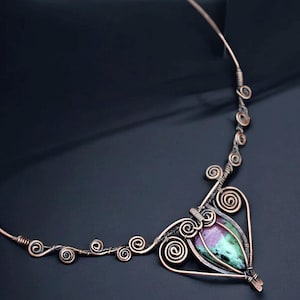 Pode incluir: Colar de arame de cobre com um pendente central com uma pedra preciosa com tons roxos e verdes. O design do colar incorpora detalhes em espiral e redemoinho, criando um visual artístico e artesanal. O colar é exibido em um fundo escuro.