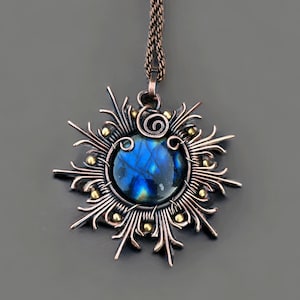May include: A handcrafted pendant necklace featuring a round, iridescent blue gemstone at the center. The stone is encased in a copper-colored metal frame with a sunburst design, accented with small gold-colored beads. The necklace hangs from a matching copper chain.