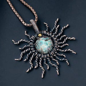 May include: A handcrafted pendant necklace featuring a sunburst design. The pendant has a central turquoise stone, surrounded by silver-toned wire rays. The necklace chain is a matching silver color. The overall design is intricate and artistic.