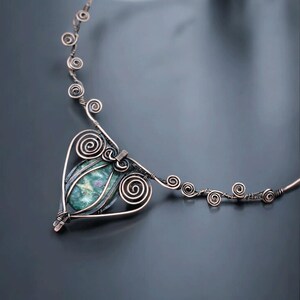 May include: A handcrafted copper wire necklace featuring a heart-shaped pendant with a green stone. The necklace has intricate spiral and swirl designs, with a delicate chain. The jewelry is displayed on a dark gray background.