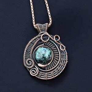 May include: A handcrafted copper pendant with a wire-wrapped design. The pendant features a central, round, green stone and intricate wire detailing. The necklace chain is also copper-colored, and the background is a dark blue.