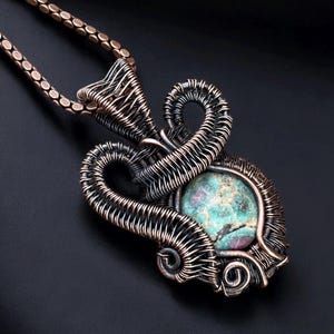 May include: Handmade copper wire-wrapped pendant necklace featuring a round, speckled turquoise and red stone. The pendant has an intricate, openwork design with a matching copper chain. The necklace is displayed against a dark background.