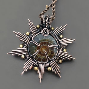 Może przedstawiać: Ręcznie robiony wisiorek z skamieniałym amonitem osadzonym w miedzianym drucianym wzorze słońca. Wisiorek zawiera miedziane druciane promienie, mosiężne koraliki i skręcony miedziany łańcuszek. Widoczny jest spiralny wzór skamieniałości.