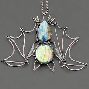 May include: A handcrafted bat pendant necklace. The pendant features a wire-wrapped design with a teardrop-shaped and a round labradorite stone. The wings and body are made of silver-colored wire, and the necklace has a silver chain.