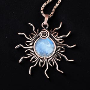 May include: A handcrafted pendant necklace featuring a sunburst design. The pendant has a central, round, light blue stone, surrounded by a copper-colored metal sunburst. The necklace chain is also copper-colored, with a circular link pattern. The background is black.