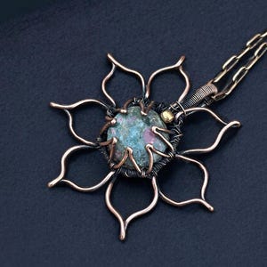 May include: A handcrafted copper pendant necklace featuring a flower design. The pendant has a central stone with blue and pink hues, surrounded by copper wire and petal-shaped outlines. The chain is a delicate gold color.