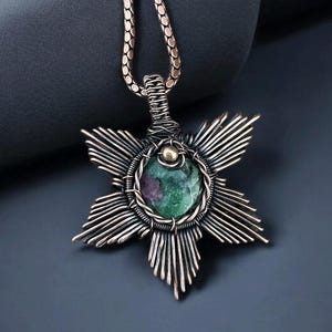 May include: A handcrafted copper pendant necklace featuring a starburst design. The pendant has a central, round, green and purple gemstone, encircled by wire. The necklace chain is a matching copper color.