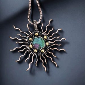May include: A handcrafted pendant necklace featuring a sunburst design. The pendant has a central, multi-faceted green and purple gemstone, surrounded by silver-toned metal rays and gold-colored accents. The necklace chain is a silver-toned, textured cable.
